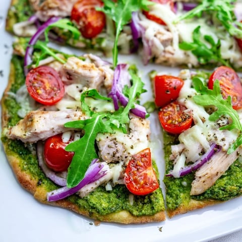A close-up of pesto chicken flatbread with bubbling cheese, fresh arugula, and tomatoes.